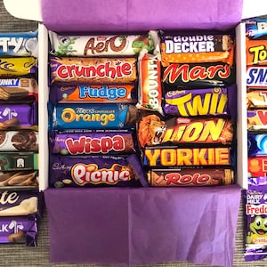 May include: A selection of chocolate bars in a box, including brands like Bounty, Kit Kat, Mars, Snickers, Twix, and others. The box is lined with purple paper, showcasing a variety of popular chocolate treats.