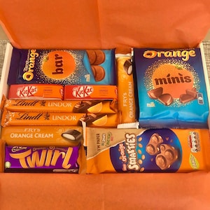 May include: A gift box filled with orange-flavored chocolate treats. The box includes Terry's Chocolate Orange, Kit Kat, Lindt Lindor, Fry's Orange Cream, and Smarties. The box is lined with orange tissue paper, creating a vibrant presentation.