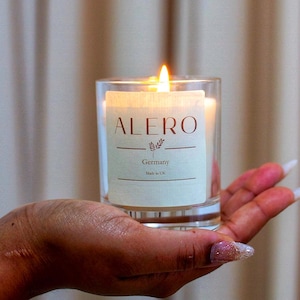 Könnte beinhalten: Eine brennende Kerze in einem durchsichtigen Glasbehälter. Auf dem Etikett der Kerze steht "ALERO Germany Made in UK". Die Kerze wird in einer Hand mit langen Fingernägeln gehalten. Der Hintergrund ist ein beiger Vorhang.
