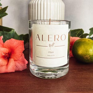 Puede incluir: Una vela de cristal transparente con una etiqueta que dice "ALERO Haiti Made in UK". La vela está sobre una superficie de madera, junto a una lima y una flor de color coral. Un jarrón blanco con follaje seco está en el fondo.