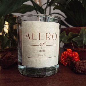 Op de afbeelding: Een heldere glazen kandelaar met een crèmekleurig etiket. Het etiket bevat het woord "ALERO" in koperkleur, met "India" en "Made in UK" eronder. De kaars staat op een houten oppervlak, met groene planten en oranje bloemen op de achtergrond.