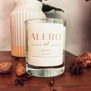 Può includere: Una candela di vetro trasparente con un'etichetta bianca con la parola "ALERO" in rame, con il testo "Australia" e "Made in UK". La candela è su una superficie di legno con spezie e un vaso bianco sullo sfondo.