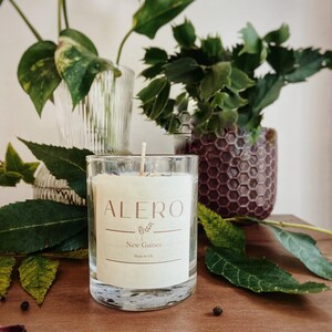 Op de afbeelding: Een heldere glazen kaars met een crèmekleurig etiket met de tekst "ALERO", "New Guinea" en "Made in UK". De kaars staat op een houten oppervlak, omringd door groene planten en gedroogde rozenblaadjes.