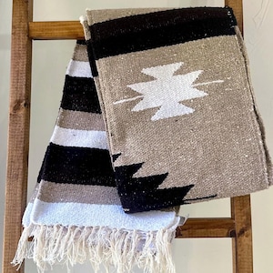 May include: A woven blanket with a geometric design in black, white, and tan. The blanket features a central white design and fringe along one edge. The blanket is draped over a wooden ladder.