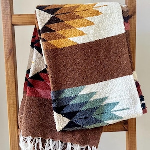 May include: A woven blanket with a brown base and geometric patterns in white, yellow, black, red, and blue. The blanket has a fringed edge and is draped over a wooden ladder.