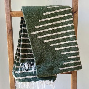 May include: A green and white striped woven blanket with fringe. The blanket is draped over a wooden ladder. The stripes are white and run horizontally across the blanket, with some angled lines.