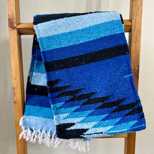 May include: A blue and black striped Mexican blanket with white fringe. The blanket is folded and draped over a wooden ladder.