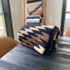 May include: Rolled woven blanket with a black background and a repeating zigzag pattern in shades of brown and cream. Other blankets with similar patterns are visible in the background, displayed on a wooden ladder.