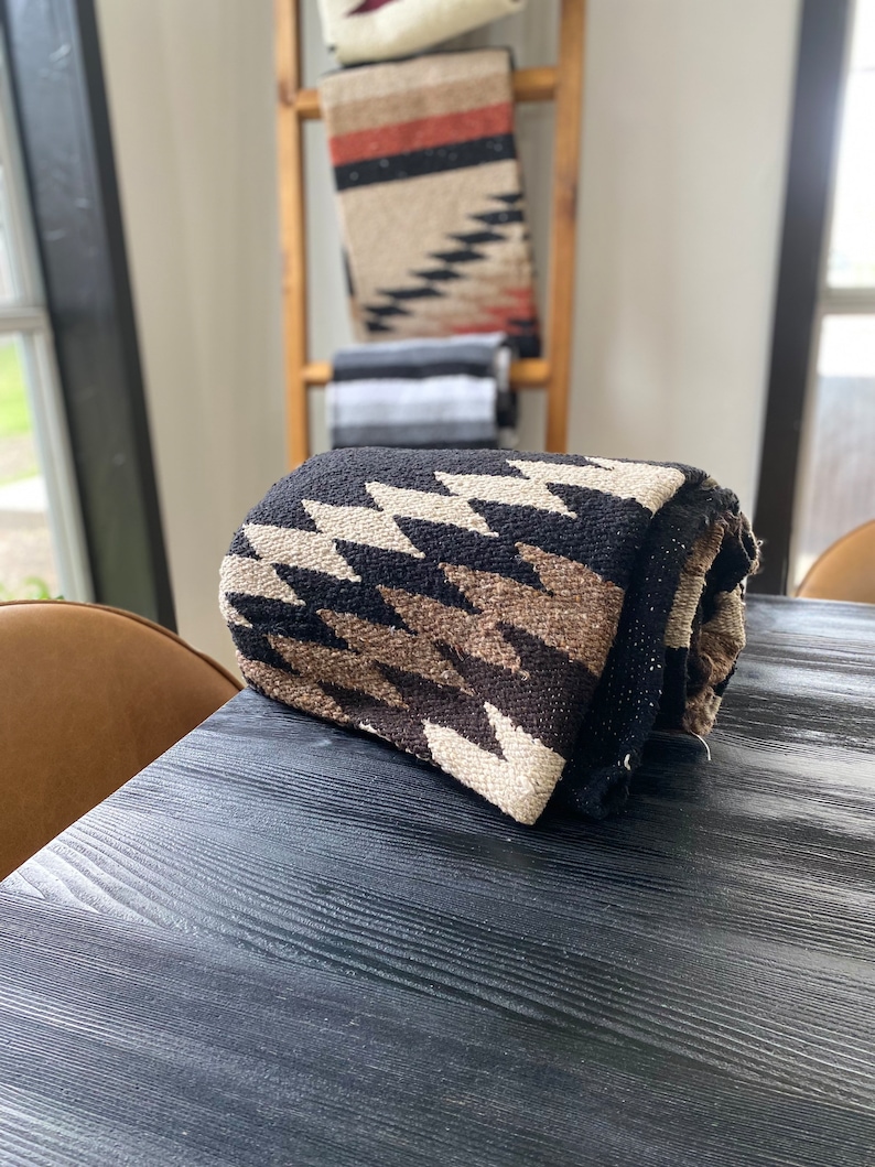 May include: Rolled woven blanket with a black background and a repeating zigzag pattern in shades of brown and cream. Other blankets with similar patterns are visible in the background, displayed on a wooden ladder.