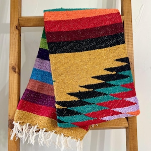 May include: A colorful woven blanket with a geometric design. The blanket features a vibrant mix of colors, including red, orange, yellow, black, green, and purple. It is draped over a wooden ladder, showcasing its texture and fringe.