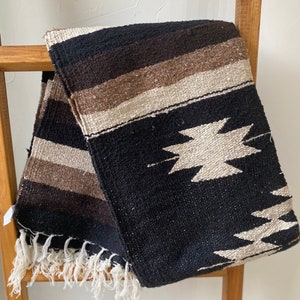 May include: A woven blanket in black, brown, and beige stripes and geometric patterns. The blanket has a fringed edge and is draped over a wooden ladder. The blanket is made of a thick, textured material.