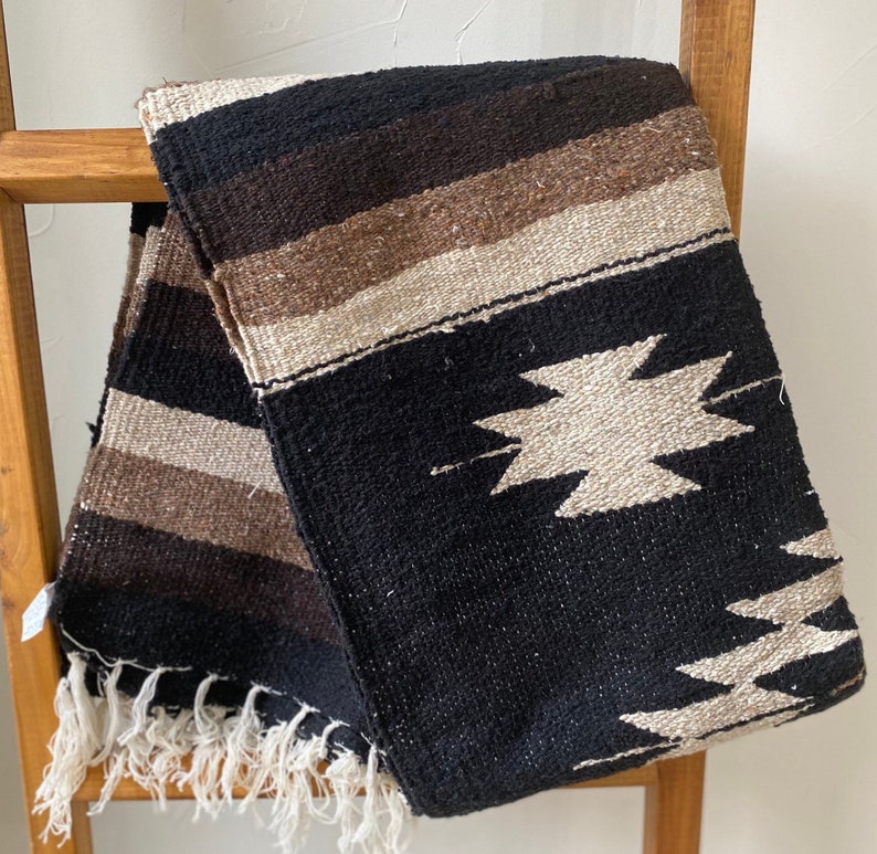 May include: A woven blanket in black, brown, and beige stripes and geometric patterns. The blanket has a fringed edge and is draped over a wooden ladder. The blanket is made of a thick, textured material.