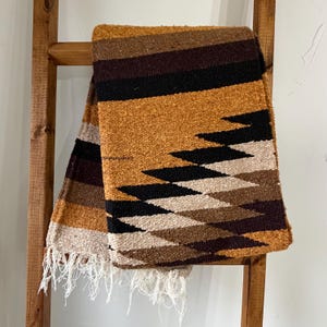 May include: A woven blanket with a geometric pattern in shades of brown, black, and beige, hanging on a wooden ladder. The blanket has a fringed edge and a textured surface, suitable for home decor.