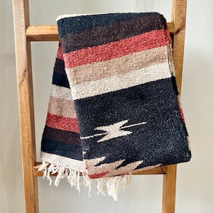 May include: A woven blanket with a striped pattern in black, brown, red, and cream colors. The blanket is draped over a wooden ladder. The bottom edge has a fringe.