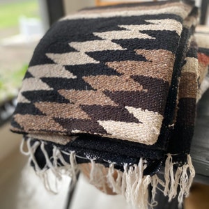 May include: Folded woven blanket with a geometric pattern in black, beige, and brown. The blanket has a fringe along the edge. The blanket is folded on a dark surface. The blanket is a southwestern style.