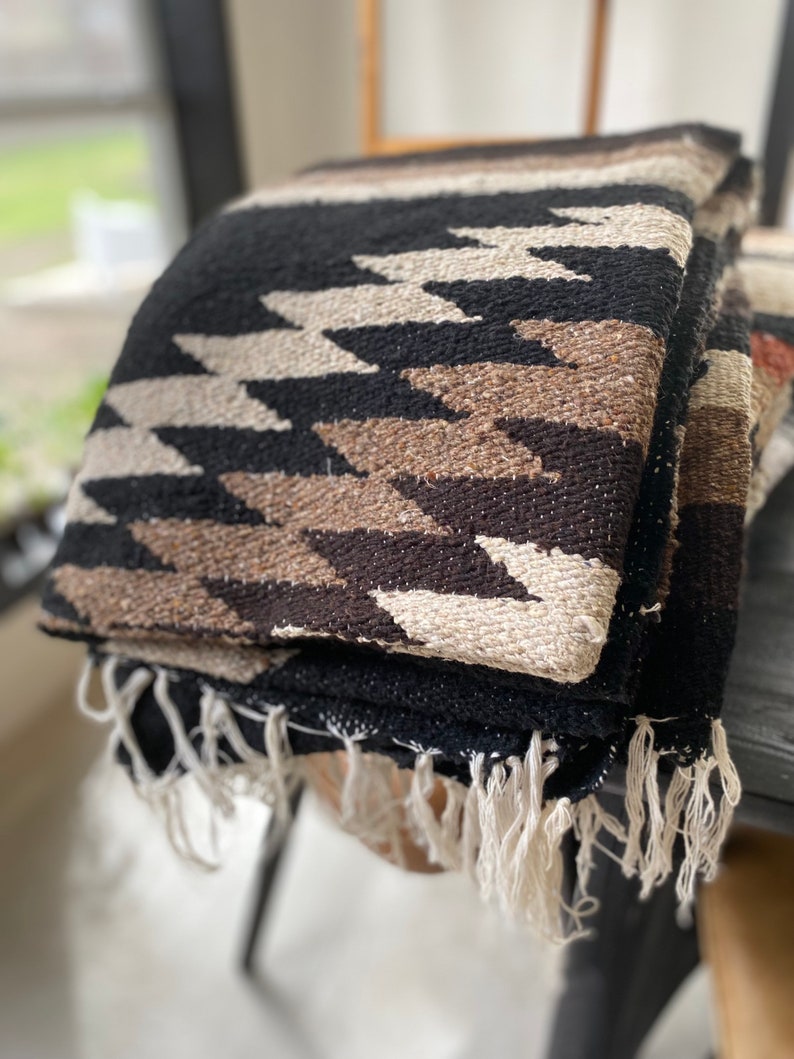 May include: Folded woven blanket with a geometric pattern in black, beige, and brown. The blanket has a fringe along the edge. The blanket is folded on a dark surface. The blanket is a southwestern style.