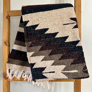 May include: A woven throw blanket with a geometric pattern in shades of brown, black, and white. The blanket has a fringe border.