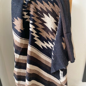 May include: A woven blanket with a geometric pattern in black, brown, and cream colors. The blanket is draped over a wooden ladder, showcasing its texture and design. The blanket features a striped border and fringed edges.