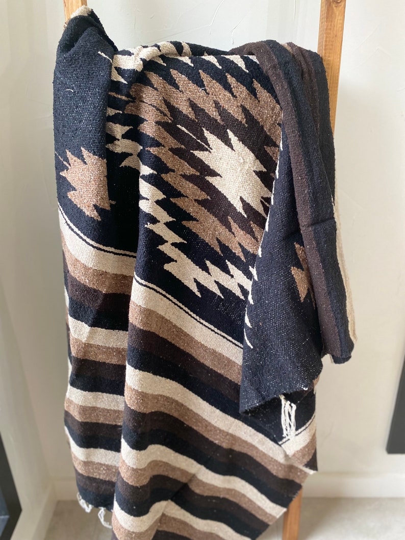 May include: A woven blanket with a geometric pattern in black, brown, and cream colors. The blanket is draped over a wooden ladder, showcasing its texture and design. The blanket features a striped border and fringed edges.