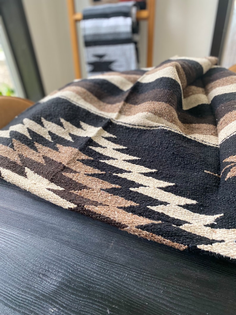 May include: A folded woven blanket with a geometric pattern. The blanket features alternating stripes of black, brown, and beige. The bottom edge has a zigzag design in black and beige. The blanket is draped over a dark surface.