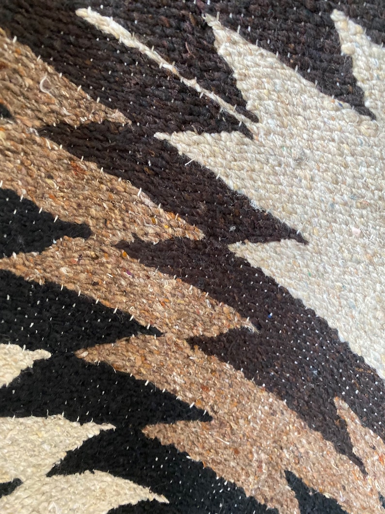 May include: Close-up of a woven textile with a geometric pattern. The design features alternating bands of black, brown, tan, and off-white, creating a zig-zag effect. The texture appears coarse and handmade, with visible stitching.