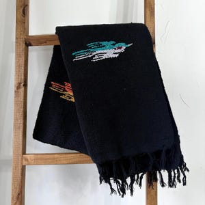 May include: Black woven blanket with a colorful embroidered design. The blanket is draped over a wooden ladder. The embroidered design features a bird-like figure in shades of teal, white, orange, and yellow. The blanket has a fringe along the bottom edge.