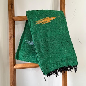 May include: A folded green blanket with a yellow and orange design, hanging on a wooden ladder. The blanket has black fringe on one end. The blanket is made of a woven material.