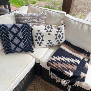 May include: An outdoor sectional sofa with cream-colored cushions and several decorative pillows. The pillows feature geometric patterns in black and white, and a gray and white design. A folded black, brown, and white blanket with fringe is also present.