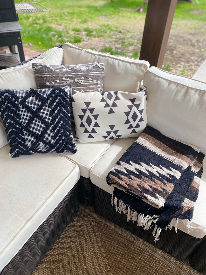 May include: An outdoor sectional sofa with cream-colored cushions and several decorative pillows. The pillows feature geometric patterns in black and white, and a gray and white design. A folded black, brown, and white blanket with fringe is also present.