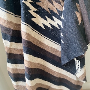 May include: A woven textile with a geometric pattern in black, brown, and cream. The design features diagonal stripes and diamond shapes. The fabric appears to be thick and textured, with fringed edges. The textile is likely a blanket or throw.