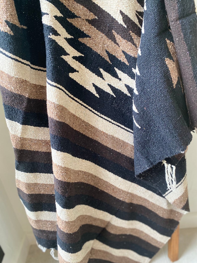 May include: A woven textile with a geometric pattern in black, brown, and cream. The design features diagonal stripes and diamond shapes. The fabric appears to be thick and textured, with fringed edges. The textile is likely a blanket or throw.