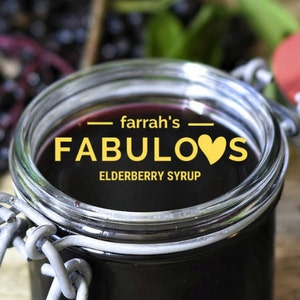 Könnte beinhalten: Ein Glasgefäß gefüllt mit dunkelviolettem Holundersaft. Das Gefäß hat einen Metallverschluss und ein Etikett mit der Aufschrift "farrah's FABULOUS ELDERBERRY SYRUP" mit einem gelben Herzen.