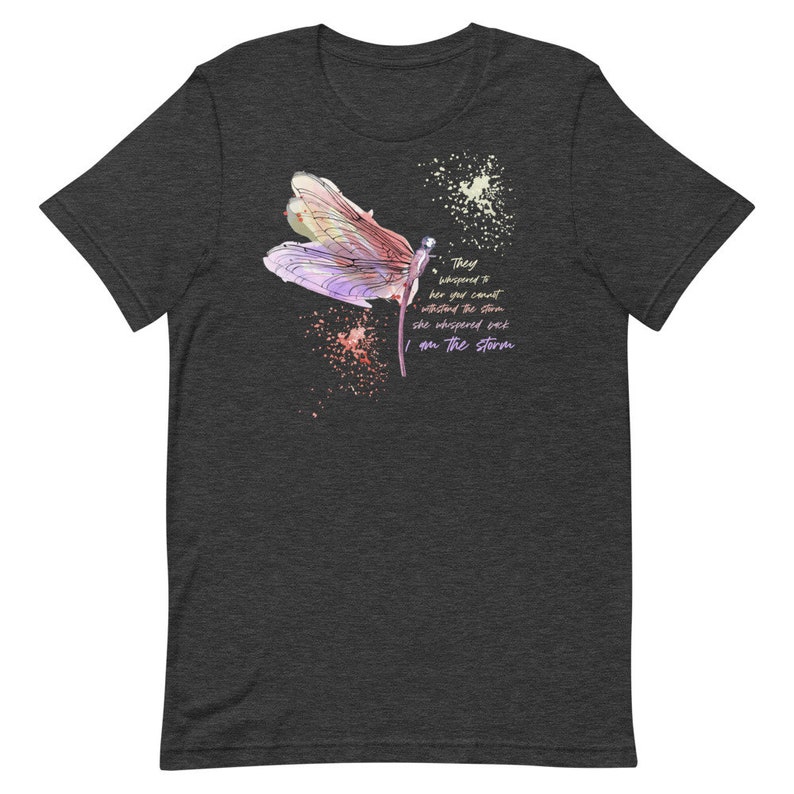May include: A dark heather grey t-shirt with a watercolor dragonfly design and the text "They whispered to her you cannot withstand the storm she whispered back I am the storm".