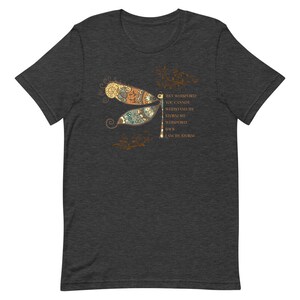 May include: Dark heather grey t-shirt featuring a dragonfly with intricate details in shades of brown, orange, and green. The dragonfly is surrounded by floral elements. The text "They whispered you cannot withstand the storm she whispered back I am the storm" is printed on the right side of the shirt.