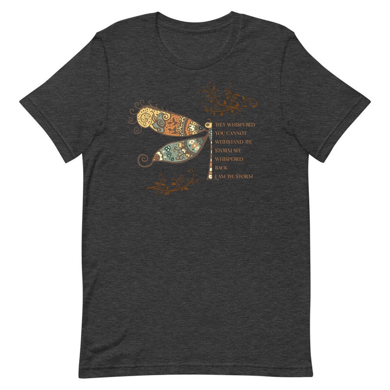 May include: Dark heather grey t-shirt featuring a dragonfly with intricate details in shades of brown, orange, and green. The dragonfly is surrounded by floral elements. The text "They whispered you cannot withstand the storm she whispered back I am the storm" is printed on the right side of the shirt.