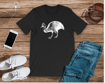 Op de afbeelding: Zwart T-shirt met een witte afbeelding van een cassowary. Het T-shirt ligt plat op een houten oppervlak met een paar witte sneakers, een blauwe jeans en een koptelefoon.