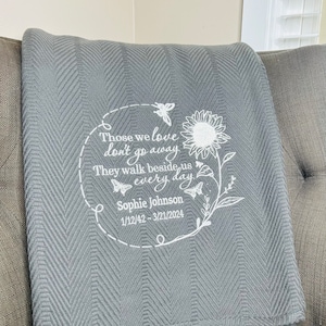 May include: Gray chevron patterned throw blanket with white embroidered text that reads "Those we love don't go away They walk beside us every day" and "Sophie Johnson 1/12/42 - 3/21/2024". The embroidery also includes a sunflower, butterflies, and leaves.