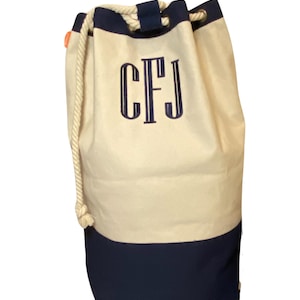 May include: A large, white canvas tote bag with a navy blue bottom. The bag has a rope drawstring closure and is personalized with the monogram "CFJ" in navy blue embroidery.