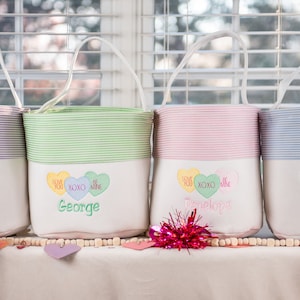 May include: Four personalized Valentine's Day gift bags with candy heart designs and names embroidered on the front. The bags are striped in different colors: purple, green, pink, and blue.