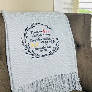 May include: A light gray herringbone throw blanket with fringe edges, featuring an embroidered wreath design in dark blue, the word love in red, and yellow angel wings. The text reads: Those we love don't go away. They walk beside us every day.