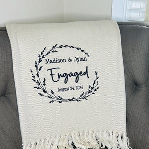 May include: A cream-colored throw blanket with a herringbone weave and fringe trim. The center features black embroidery of a leafy wreath surrounding the text Madison & Dylan, Engaged, and the date August 24, 2024, in a mix of script and serif fonts.