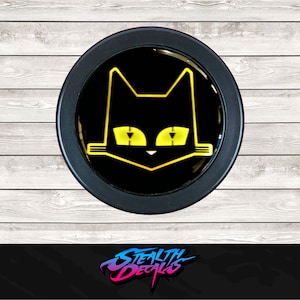 Op de afbeelding: Een zwarte en gele sticker met een kattengezicht op een zwarte ronde achtergrond. De kat heeft gele ogen en een gele neus.