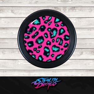 May include: A round, black object with a vibrant pink leopard print design, accented with teal and black spots. The object is set against a light wood grain background. The Stealth Decals logo is at the bottom.