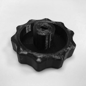 Op de afbeelding: Een zwarte plastic knop met een stervormig ontwerp en een centraal gat. De knop heeft een getextureerd oppervlak.