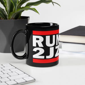 May include: Black ceramic mug with the text "RUN 2JZ" in white letters on a red stripe. The mug has a black handle and is sitting on a white surface. A plant and a book are in the background.