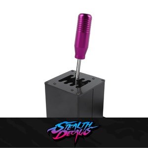 Könnte beinhalten: Ein schwarzer Metall-Schalthebel mit einem violetten Schaltknauf. Der Schalthebel hat ein 5-Gang-Schema.