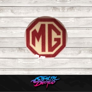 May include: A red and white MG logo decal. The logo is a red octagon with the letters MG in white inside. The decal is on a white wooden background.