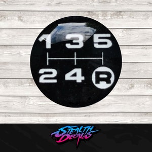 May include: A black gear shift knob with white numbers and a reverse indicator. The numbers 1, 3, 5, 2, 4, and R are displayed. The image is set against a light wood grain background.