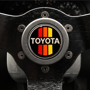 May include: A black steering wheel with a silver Toyota logo in the center. The logo has red, orange, and yellow stripes.