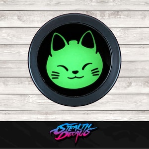 May include: A round, black decal featuring a smiling green cat face. The cat has pointed ears and three whiskers on each side. The decal is set against a light wood grain background. The text "STEALTH DECALS" is at the bottom.